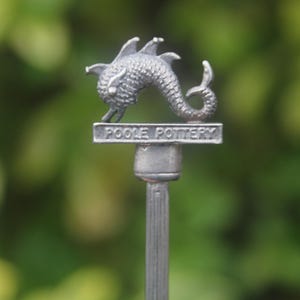 May include: A silver Poole Pottery figurine of a sea creature with a curved tail. The figurine is mounted on a slender, vertical post, with the words "POOLE POTTERY" inscribed on a rectangular base.