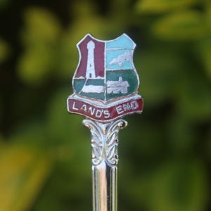 May include: A silver spoon with a decorative handle and a colorful enamel crest. The crest features a lighthouse, train, and the words "LAND'S END" in red lettering. The background is blurred green.