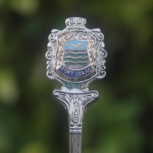 May include: A silver spoon with an ornate design, featuring a crest with blue and green details. The crest has the words "PORT ISAAC" in a blue banner. The spoon is set against a blurred green background.