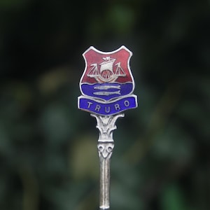 May include: A silver spoon with a decorative handle and a shield-shaped emblem. The emblem features a red and blue design with a ship, fish, and the word "TRURO" in blue lettering.