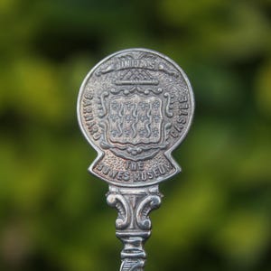 May include: A silver spoon with an ornate handle and a circular emblem. The emblem features a coat of arms and the text "BARNARD CASTLE" and "THE BOWES MUSEUM". The background is a blurred green.