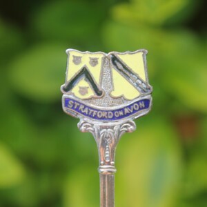 May include: A silver spoon with a decorative handle and a shield-shaped emblem. The emblem features a yellow background with black and blue details and the words "STRATFORD ON AVON" in blue.