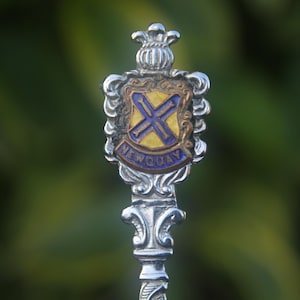 May include: A silver souvenir spoon with a blue and yellow enamel design. The spoon features a crest with a cross and the words "Newquay" on it.