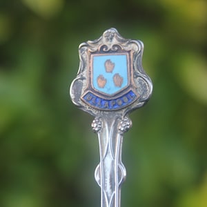 May include: A vintage silver spoon with a decorative handle and a shield-shaped emblem. The emblem features a light blue background with three brown hands and the word "ULLAPOOL" in blue lettering. The spoon is likely a souvenir.