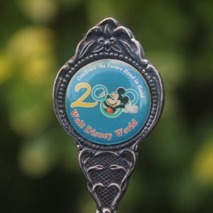Op de afbeelding: Een zilveren lepel met een turquoise embleem met Mickey Mouse en de tekst "20 Walt Disney World". Het embleem is omgeven door een decoratieve rand en de steel heeft een sierlijk ontwerp. De lepel herdenkt de 20e verjaardag.