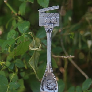 May include: A silver-coloured spoon featuring a film clapperboard design. The spoon's handle has an ornate design and a film director's chair at the base. The background is a blurred view of green leaves and foliage.