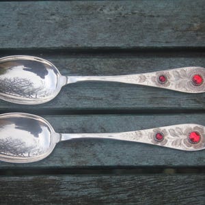 May include: Two silver spoons with ornate floral and red jewel detailing on the handles. The spoon bowls reflect a tree-lined landscape. The spoons are displayed on a weathered, teal-colored wooden surface.