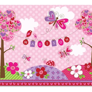 May include: Pink placemat with a heart-shaped tree design, butterflies, birds, and the name "MADISON" spelled out with hanging ornaments. The placemat has a pink and white polka dot background and a green and pink border.
