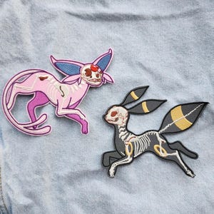 Puede incluir: Dos parches bordados con criaturas esqueléticas estilizadas. Uno es rosa con detalles morados, el otro es negro con detalles amarillos. Ambos tienen estructuras óseas detalladas y están sobre un fondo azul claro.