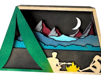 May include: A wooden layered art piece depicting a camping scene. A green tent frames a night sky with mountains, a crescent moon, and figures around a campfire. The artwork uses shades of green, blue, red, and yellow.