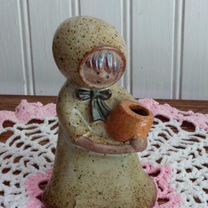 May include: A ceramic figurine of a person wearing a hooded cloak, holding a small orange pot. The figurine has a speckled, tan finish with a dark bow at the neck. The face is visible through the hood.