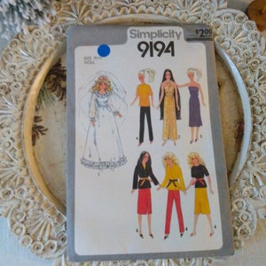 May include: Simplicity sewing pattern number 9194 for 11 1/2 inch dolls. The pattern includes five different outfits, including a wedding dress, a jumpsuit, a skirt and top, and two different pants and top combinations. The pattern is printed on a gray background with black and white illustrations.