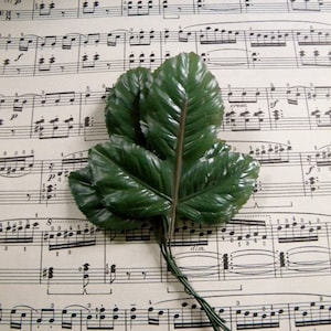 May include: A bouquet of three green artificial leaves on a sheet of music with musical notes printed in black. The leaves are arranged in a fan shape and have a wire stem.