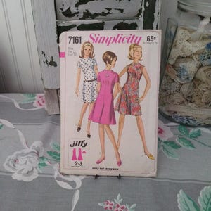 May include: A Simplicity 7161 sewing pattern envelope featuring illustrations of three different dress styles. The cover displays the text 7161, Simplicity, 65c, Miss size 12, bust 32, and Jiffy 2-3 easy cut-easy sew. The dresses are white with black dots, solid pink, and a floral print.