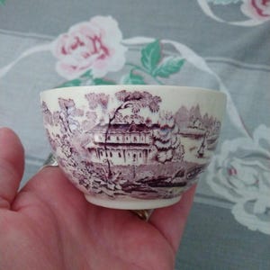 May include: A cream-colored ceramic teacup featuring a detailed dark purple transferware pattern. The design depicts a landscape with a house, trees, and a body of water. The cup has a smooth, rounded shape and a small base, showcasing a classic aesthetic.