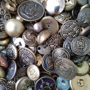 May include: A collection of vintage buttons in various shapes, sizes, and metallic finishes. The buttons are primarily gold, silver, and bronze, with intricate embossed designs, including floral patterns, crests, and geometric shapes. Some buttons have a domed or flat appearance.