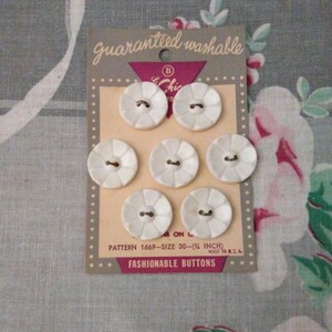 May include: A card of seven white, round, scalloped-edge buttons with two holes in the center. The packaging is gray and cream with the text "guaranteed washable," "Le Chic," "PATTERN 1669-SIZE 30-(3/4 INCH)," and "FASHIONABLE BUTTONS" printed on it.
