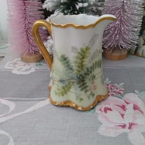 May include: A small, white ceramic pitcher with a gold handle and trim. The pitcher is decorated with green fern-like leaves and small pink flowers. The pitcher is approximately 5 inches tall.