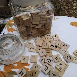 May include: A glass jar filled with wooden letter tiles, with some tiles scattered on a white and brown patterned surface. The tiles are from a Scrabble game and include letters like A, E, I, O, U, and others.