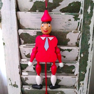 May include: A wooden marionette doll with a red body, arms, and legs, and a red pointed hat. The doll has a painted face with a smiling expression, a white collar, and a blue tie. The doll is hanging from a red string.