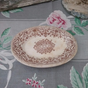 May include: Oval-shaped antique serving dish with a brown floral pattern on a cream background. The dish has a decorative border and a central floral medallion. The dish is placed on a floral patterned tablecloth.