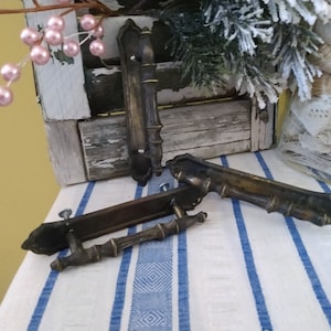 May include: Three antique brass door handles. Two are long and rectangular with ornate details. The third is a smaller, more traditional style with a rounded top.