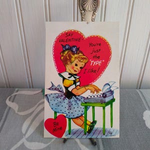 May include: Vintage Valentine's Day card featuring a cartoon girl typing on a typewriter. The card has a red heart with the text "Say Valentine - You're just the 'TYPE' I like!" and a smaller heart that says "Be Mine."