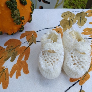 May include: A pair of white crocheted baby booties with lace trim. The booties are on a white tablecloth with a brown and orange leaf pattern.