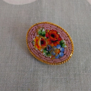 May include: Oval mosaic brooch with a floral design. The brooch features a gold-tone border and a pink background. The mosaic includes orange, red, yellow, blue, and green flowers. A vintage-style accessory.