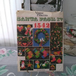 May include: Vintage Vogue Patterns Santa Sack IV pattern packet. The cover features colorful illustrations of Christmas ornaments, stockings, and a wreath. The pattern number is 1542. The packet is displayed on a floral patterned tablecloth.