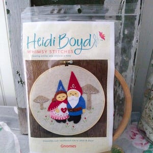 May include: A needlework kit to stitch and share featuring a pair of gnomes. The kit includes a felt gnome couple, embroidery floss, and a wooden embroidery hoop. The gnomes are dressed in red, blue, and white. The kit is packaged in a clear plastic bag with the text "Heidi Boyd Whimsy Stitches Sewing smiles one stitch at a time" and "Irresistibly cute needlework kits to stitch & share Gnomes".