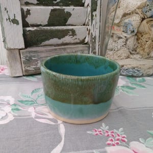 May include: A handmade ceramic bowl with a turquoise interior and a two-tone exterior. The top half is a speckled olive green, and the bottom half is a light turquoise. The bowl sits on a cream-colored base.
