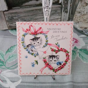 May include: A vintage Valentine's Day card with two kittens inside heart-shaped floral wreaths. The card reads "VALENTINE GREETINGS to my Teacher" in script. Pink ribbons and small blue flowers decorate the card.