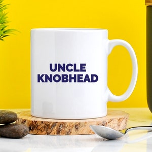 May include: White ceramic mug with a blue inscription that reads "UNCLE KNOBHEAD". The mug is sitting on a wooden coaster.
