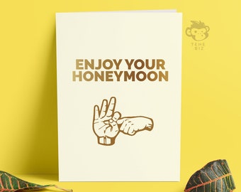 Peut inclure: Une carte blanche avec du texte doré qui dit "Enjoy your honeymoon" et une illustration dorée de deux mains faisant un geste suggestif.