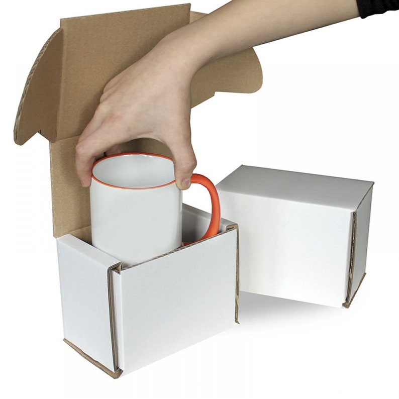 May include: A white ceramic mug with an orange handle is being placed into a white cardboard box. A second white cardboard box is visible to the right.