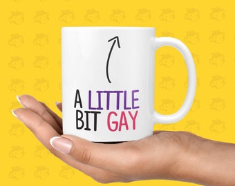 May include: White ceramic mug with a black arrow pointing upwards and the text "A LITTLE BIT GAY" in purple and pink lettering.
