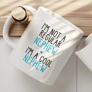 May include: A white ceramic mug featuring the black and blue text I'M NOT A REGULAR NEPHEW I'M A COOL NEPHEW with a small blue star icon between the two lines of text. The mug has a curved handle and a smooth, glossy finish on the exterior surface.