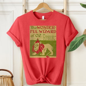 Short sleeve heather red shirt with vintage book cover of The Wizard of Oz in pale gold with grass green and red font & the cowardly lion wearing glasses. The lion's mane is red and his glasses are green. The lion is set on a light green background.