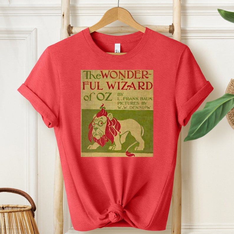 Short sleeve heather red shirt with vintage book cover of The Wizard of Oz in pale gold with grass green and red font & the cowardly lion wearing glasses. The lion's mane is red and his glasses are green. The lion is set on a light green background.