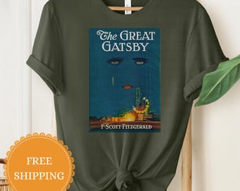 Könnte beinhalten: Ein olivgrünes T-Shirt mit Rundhalsausschnitt und einem Grafikdruck des Buchcovers von The Great Gatsby. Das Design zeigt einen blauen Hintergrund mit stilisierten Augen, roten Lippen und einer Skyline sowie den Text The Great Gatsby und F. Scott Fitzgerald.
