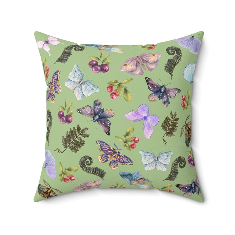 Square pillow covered with moths and butterflies in shades of blue and purple interspersed with clusters of blue berries, red berries, and fern fronds, all on a a light green background. Design on both sides of pillow