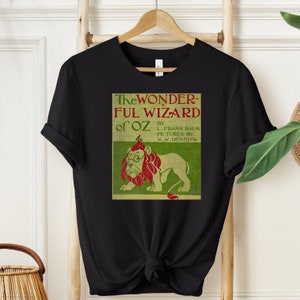 Short sleeve black shirt featuring vintage book cover of The Wizard of Oz in pale gold with grass green and red font & the cowardly lion wearing glasses. The lion's mane is red and his glasses are green. The lion is set on a light green background.