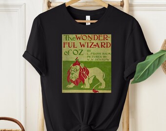 Może przedstawiać: Czarny T-shirt z vintage ilustracją tchórzliwego Lwa z "Cudownego Czarodzieja z Krainy Oz" autorstwa L. Franka Bauma. Ilustracja jest w kolorze zielonym, czerwonym i żółtym.