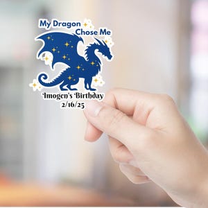 May include: A blue dragon with gold stars on its wings and body. The dragon is holding a banner that says "My Dragon Chose Me" and "Imogen's Birthday 2/16/25".