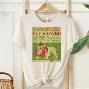 Short sleeve shirt in natural with vintage book cover of The Wizard of Oz in pale gold with grass green and red font & the cowardly lion wearing glasses. The lion's mane is red and his glasses are green. The lion is set on a light green background.