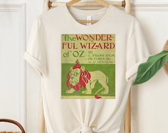Może przedstawiać: Biały T-shirt z vintage'owym wzorem okładki książki przedstawiającym lwa z czerwoną grzywą i koroną. Tekst na okładce brzmi "The Wonderful Wizard of Oz" autorstwa L. Franka Bauma, Ilustracje W.W. Denslowa.