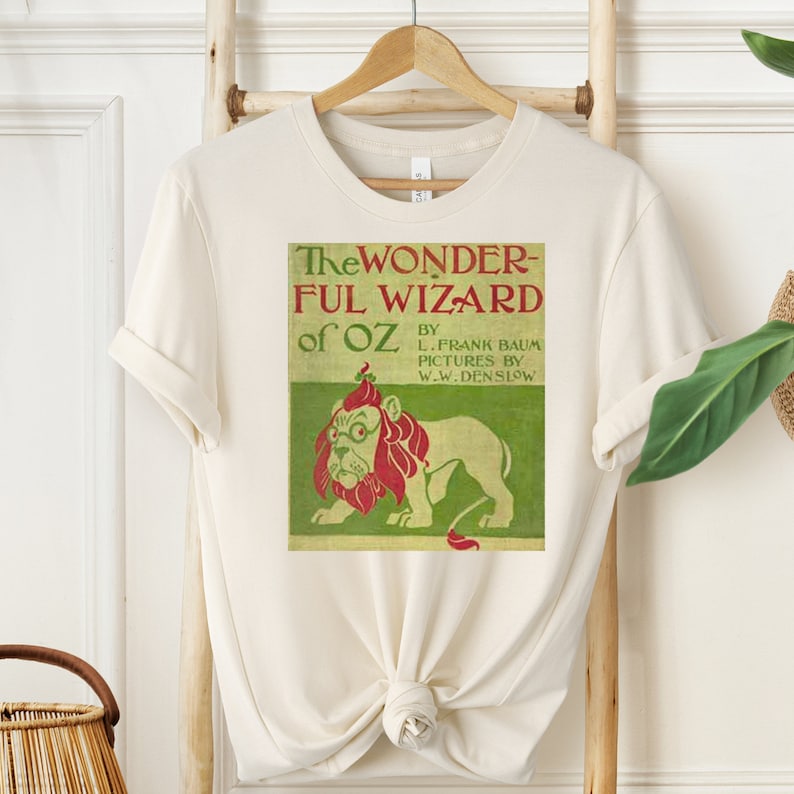 Short sleeve shirt in natural with vintage book cover of The Wizard of Oz in pale gold with grass green and red font & the cowardly lion wearing glasses. The lion's mane is red and his glasses are green. The lion is set on a light green background.