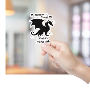 May include: A black dragon sticker with the text "My Dragon Chose Me" and "Violet's Sweet 16th". The dragon has large wings and is surrounded by small star-shaped accents. The sticker is held by a hand.
