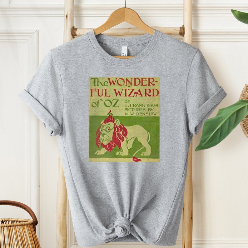 Short sleeve heather grey shirt with vintage book cover of The Wizard of Oz in pale gold with grass green and red font & the cowardly lion wearing glasses. The lion's mane is red and his glasses are green. The lion is set on a light green background.